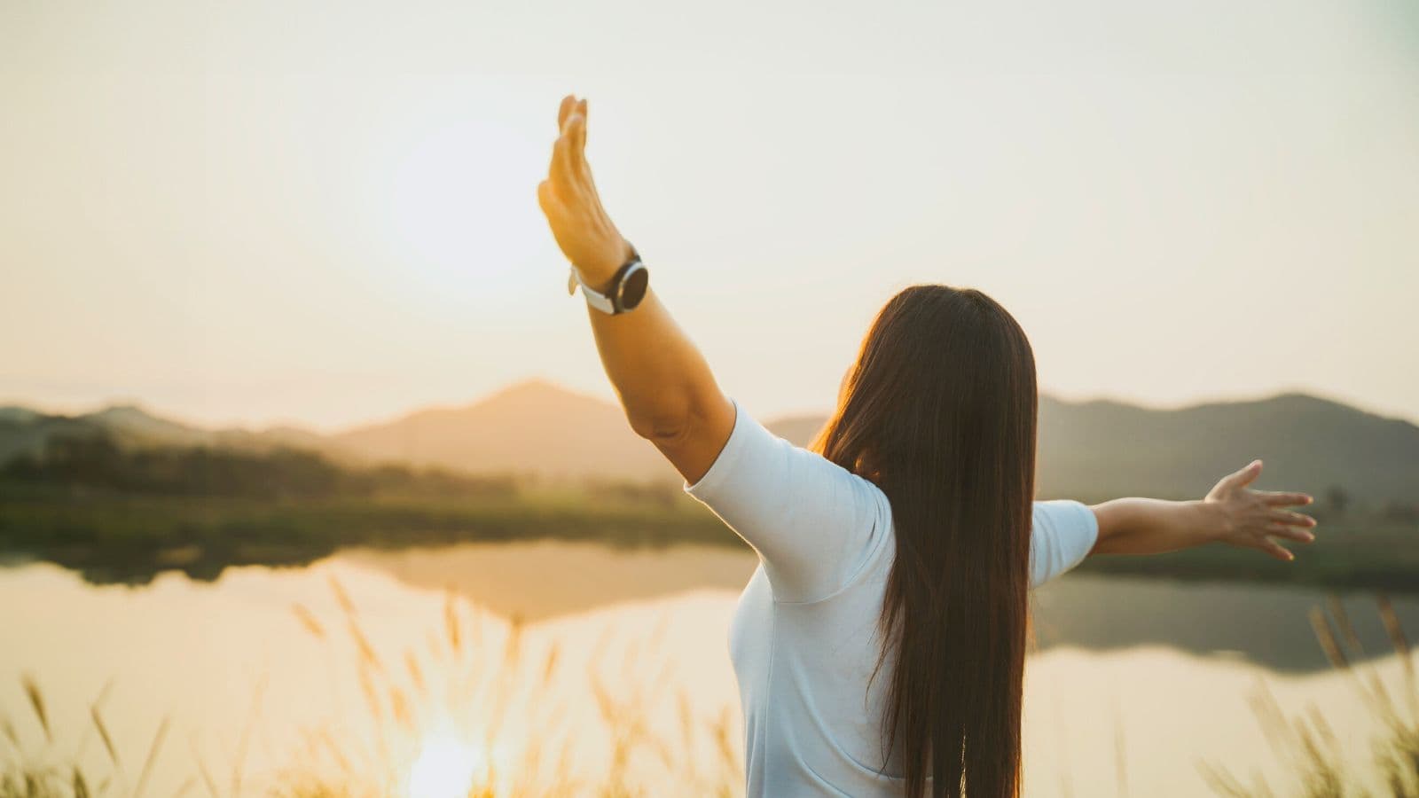 A person with long hair stands outdoors facing a lake and mountains at sunset, with arms outstretched. The individual wears a white shirt and a watch, and tall grass is visible in the foreground.