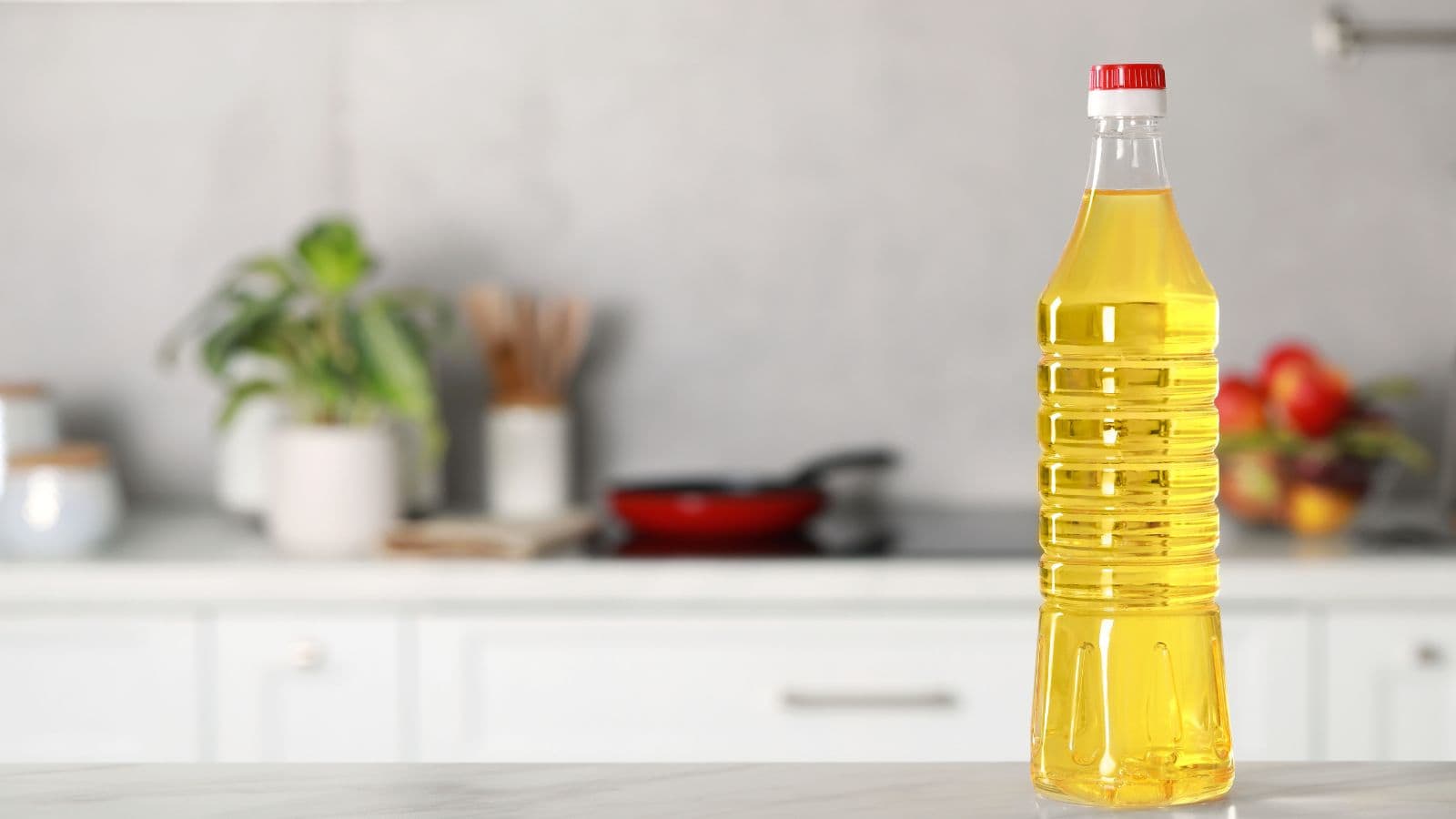 A clear plastic bottle filled with yellow cooking oil sits on a kitchen counter. The background features blurred kitchen items, including a plant, utensils, and a skillet.