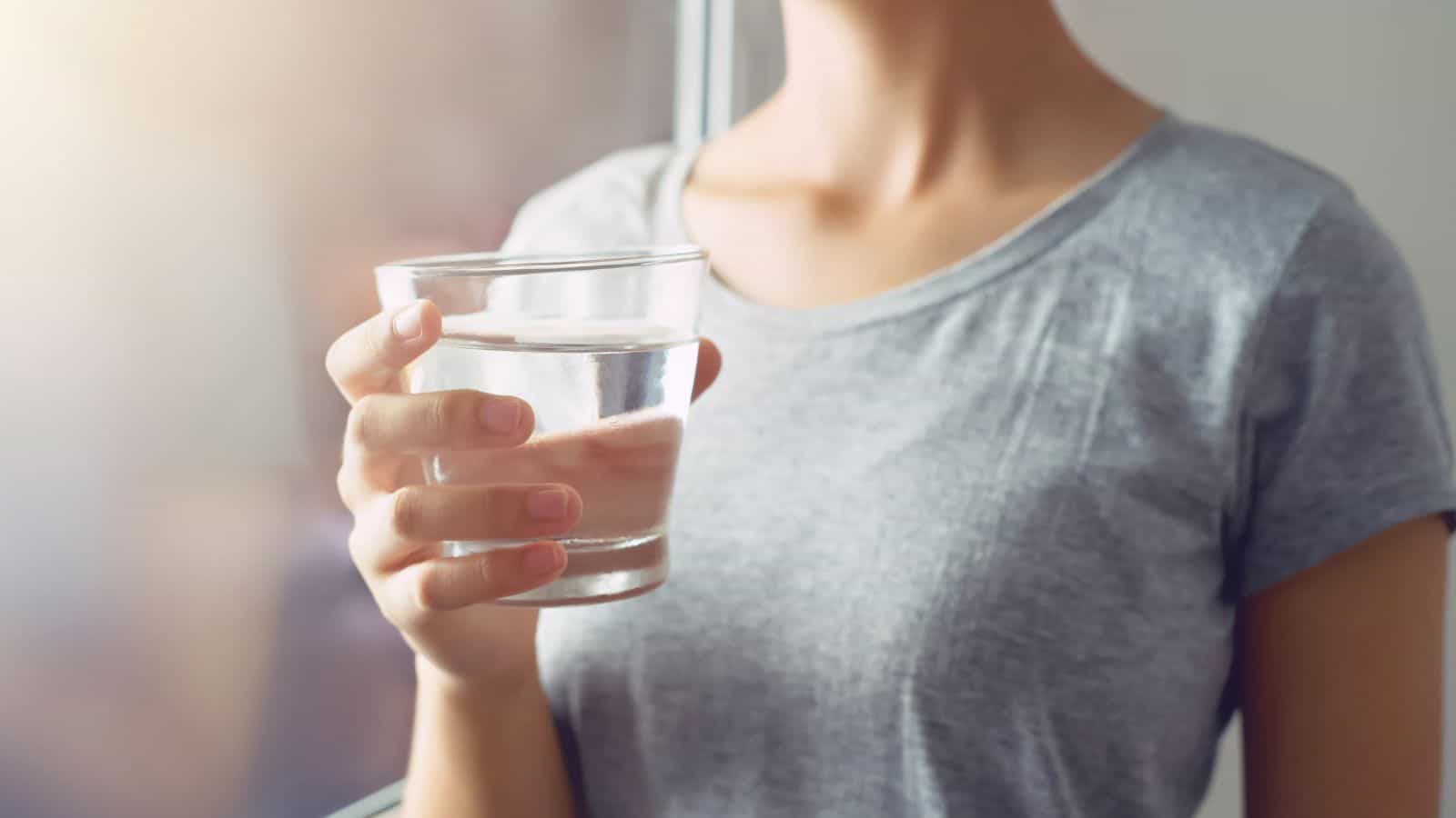 A person wearing a grey t-shirt holds a clear glass of water in their right hand while standing by a window with soft natural light. The person's face is not visible.