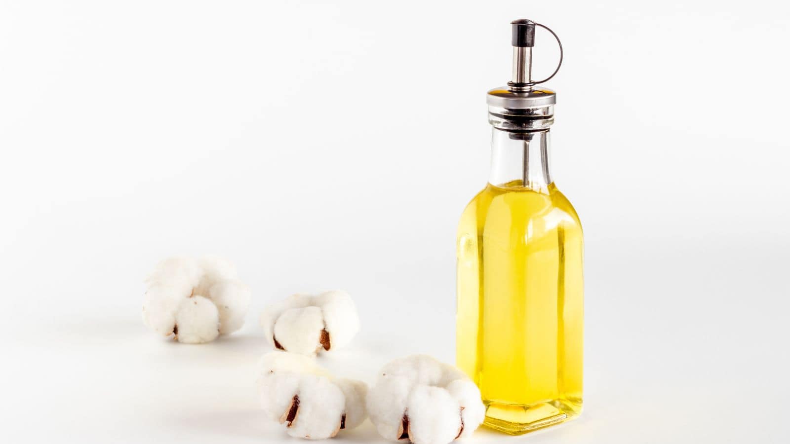 A clear glass bottle filled with yellow cottonseed oil sits on a white surface next to three pieces of raw cotton with visible brown seeds.