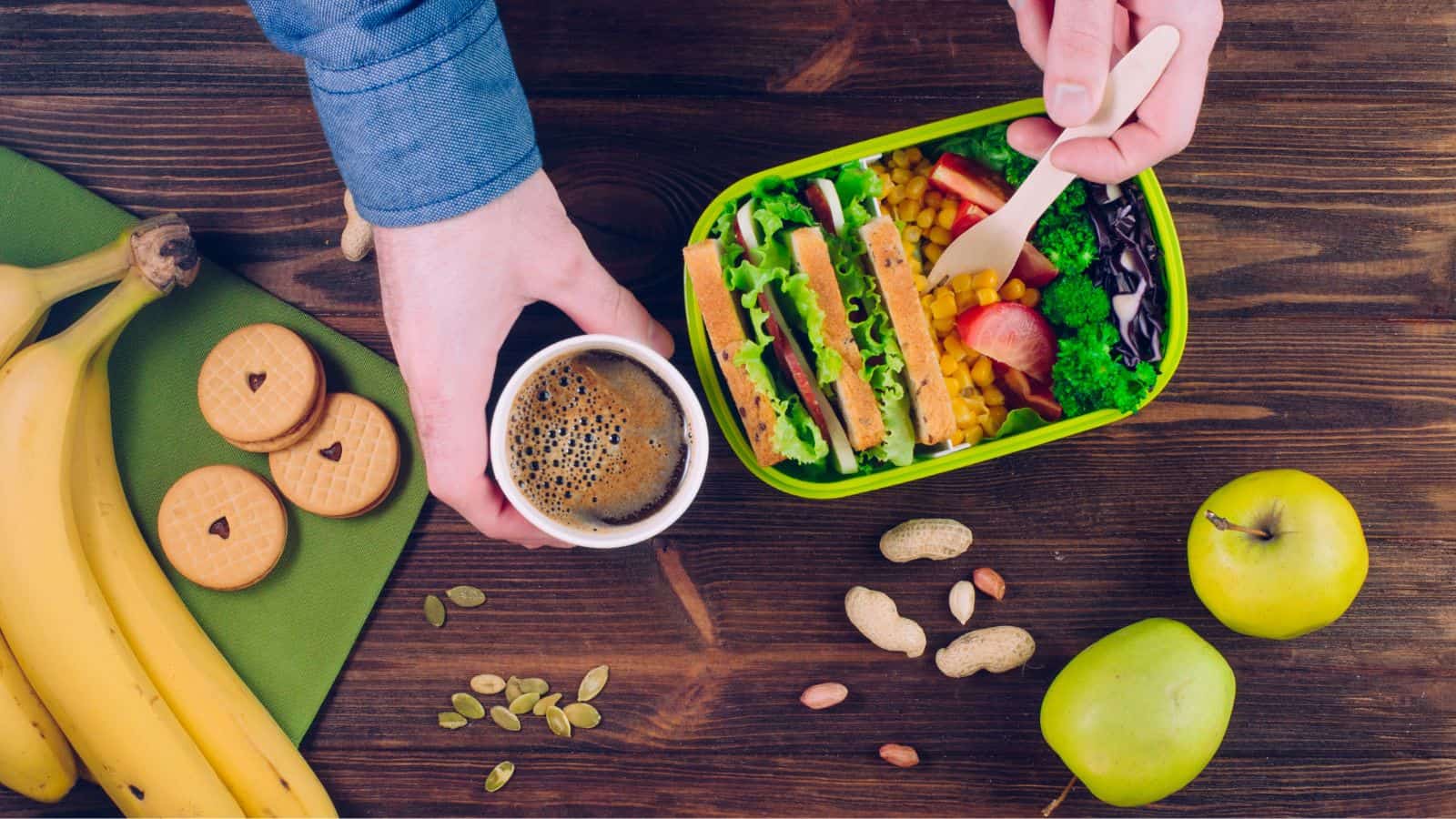 A person holds a cup of coffee in one hand and uses a wooden fork to eat from a lunchbox containing salad, sandwich, corn, and fruit. Nearby are bananas, cookies, peanuts, and green apples on a wooden table.