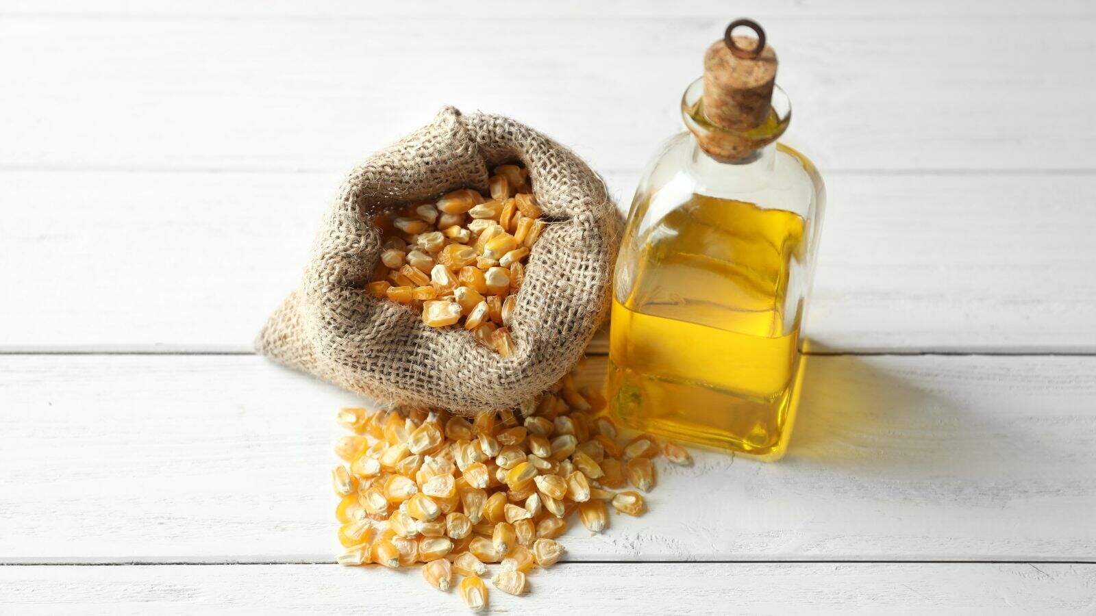 A glass bottle of corn oil with a cork top stands next to a burlap sack spilling raw corn kernels onto a white wooden surface.