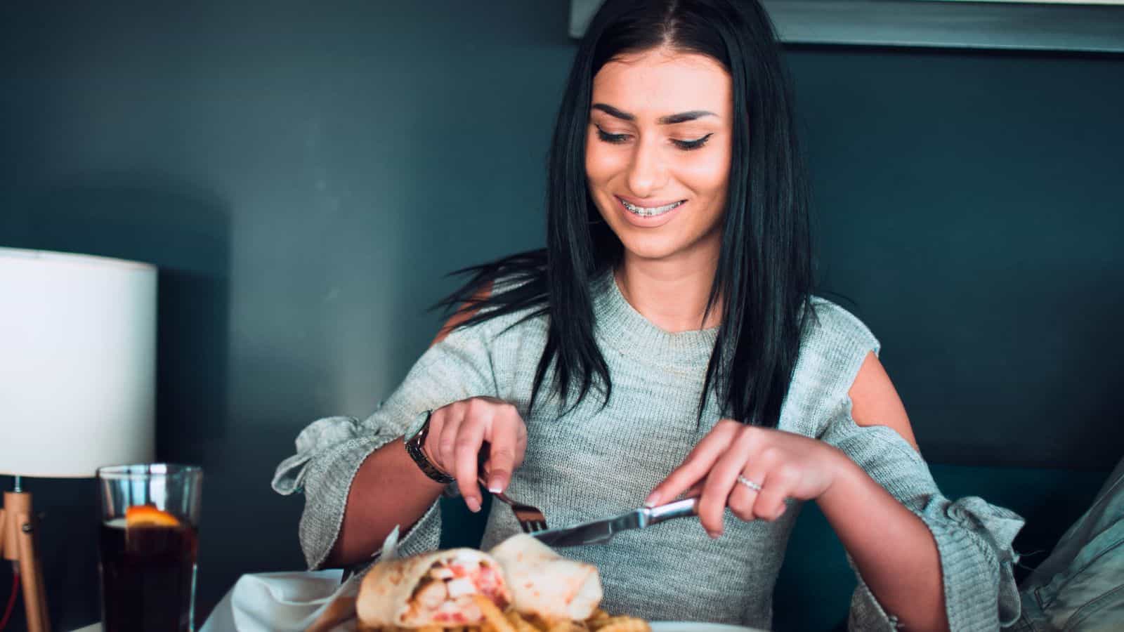 A woman with long dark hair smiles while cutting into a wrap on a plate of food. A drink with a lemon slice sits on the table beside her. She is wearing a light gray sweater and sitting indoors.