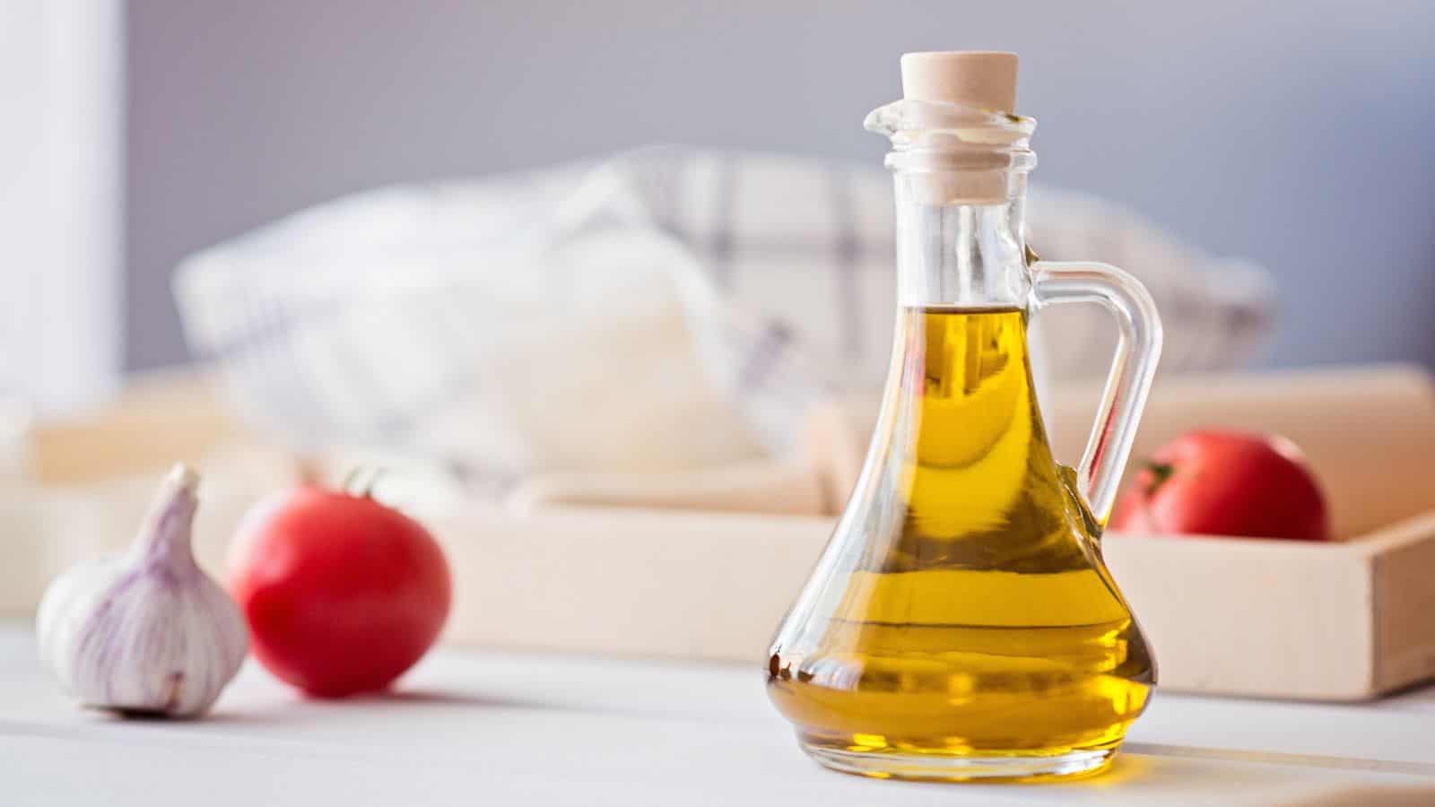 A glass bottle filled with yellow oil, sealed with a wooden stopper, is placed on a white surface. Nearby are a garlic bulb, a red tomato, and a second tomato with a wooden tray and checkered cloth in the background.