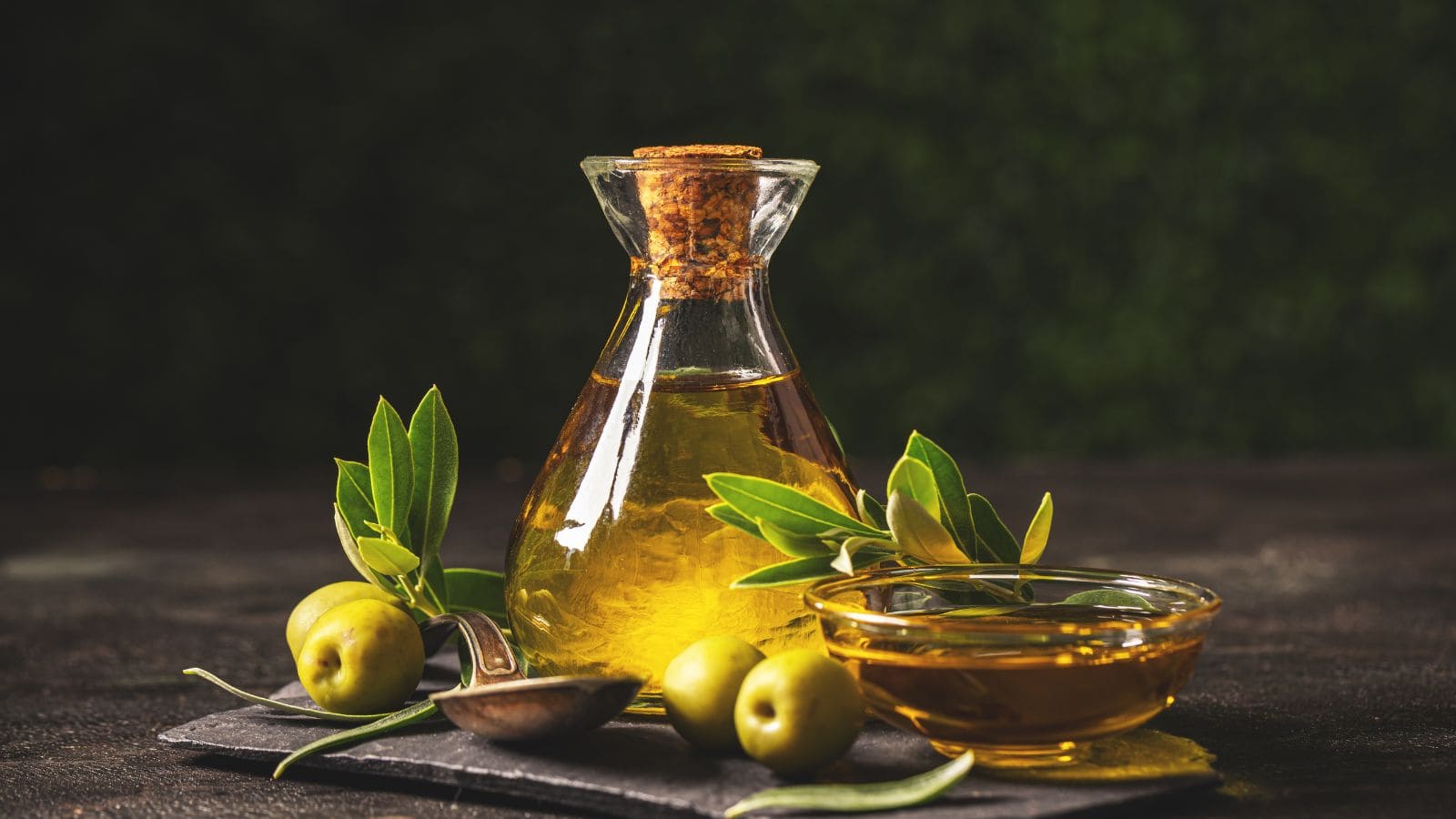 A glass bottle filled with olive oil and sealed with a cork sits next to a small glass bowl of olive oil, fresh green olives, olive branches, and a metal spoon on a dark surface with a dark background.