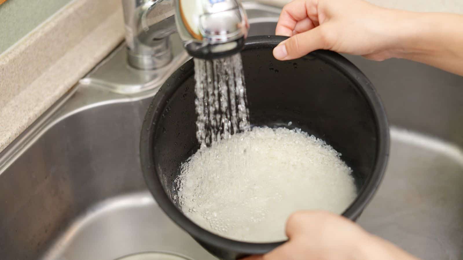 Hands holding a black pot under a kitchen faucet while rinsing white rice; water is flowing into the pot, and the rice water appears cloudy.