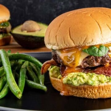 A cheeseburger with lettuce, bacon, melted cheese, and guacamole on a bun sits on a black plate next to a serving of cooked green beans. Another burger and half an avocado are in the background.