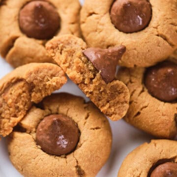 A plate of peanut butter cookies topped with chocolate kisses. One cookie is broken in half, showing a soft, crumbly texture inside. Other whole cookies surround it on the plate.