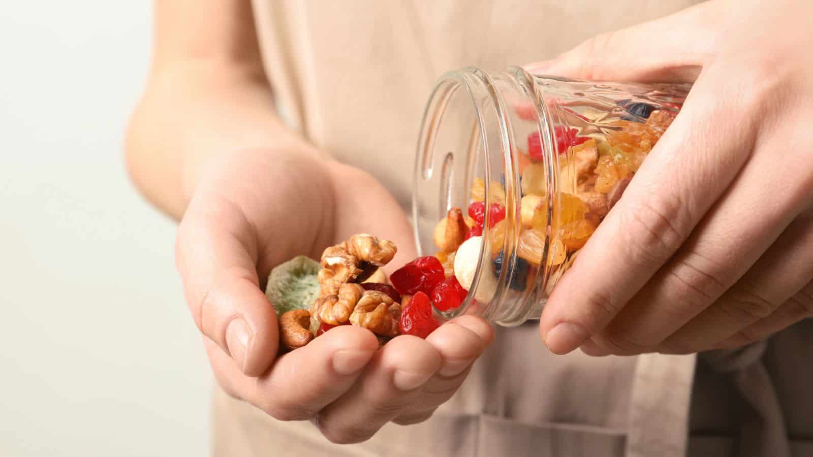 A person pours a mixture of dried fruits and nuts from a glass jar into their hand. The mix includes walnuts, dried cherries, raisins, and other dried fruits. The background is neutral and out of focus.
