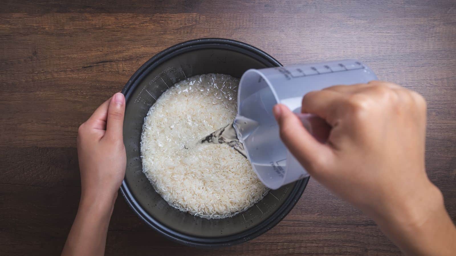 A person pours water from a measuring cup into a bowl of uncooked rice, set on a wooden surface.