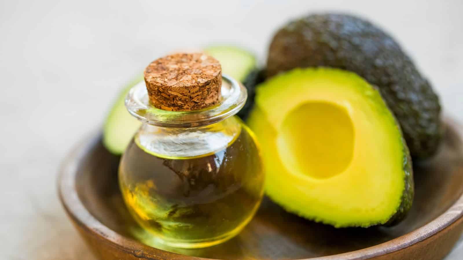 A round wooden bowl with a glass bottle of avocado oil and a cork stopper, next to a halved avocado and an uncut avocado in the background.
