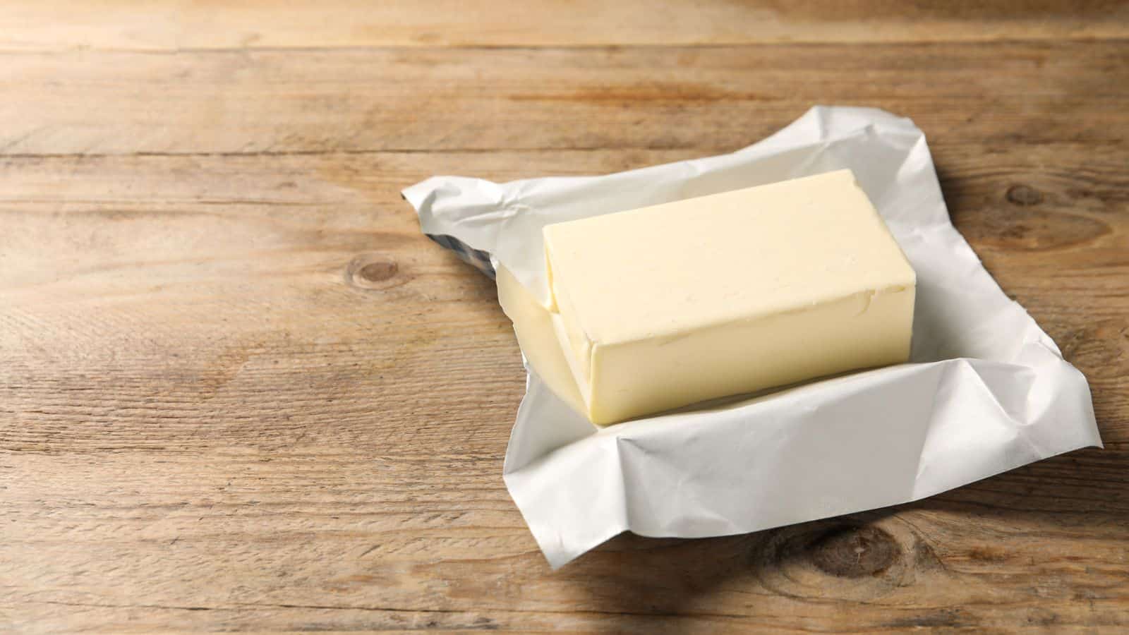 A rectangular block of butter sits on an opened wrapper on a wooden surface. The butter appears unused and the paper is slightly crumpled around it.