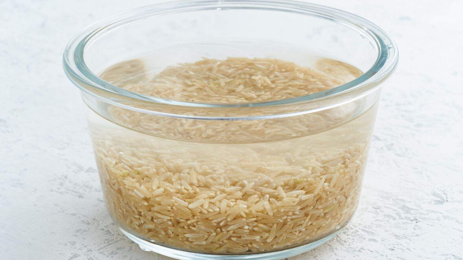A glass bowl filled with uncooked brown rice soaking in water, placed on a white surface.