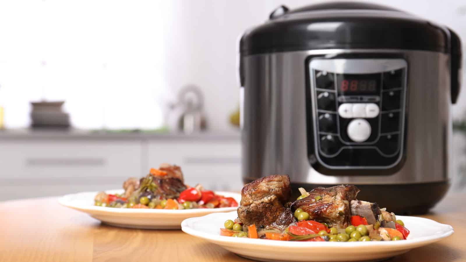 Two plates of cooked meat with vegetables are placed on a wooden table in front of a digital pressure cooker in a kitchen setting. The pressure cooker display shows numbers and buttons.