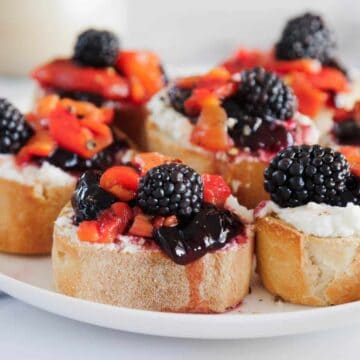 A plate of sliced baguette topped with ricotta cheese, blackberry jam, diced strawberries, and fresh blackberries, arranged on a white surface.