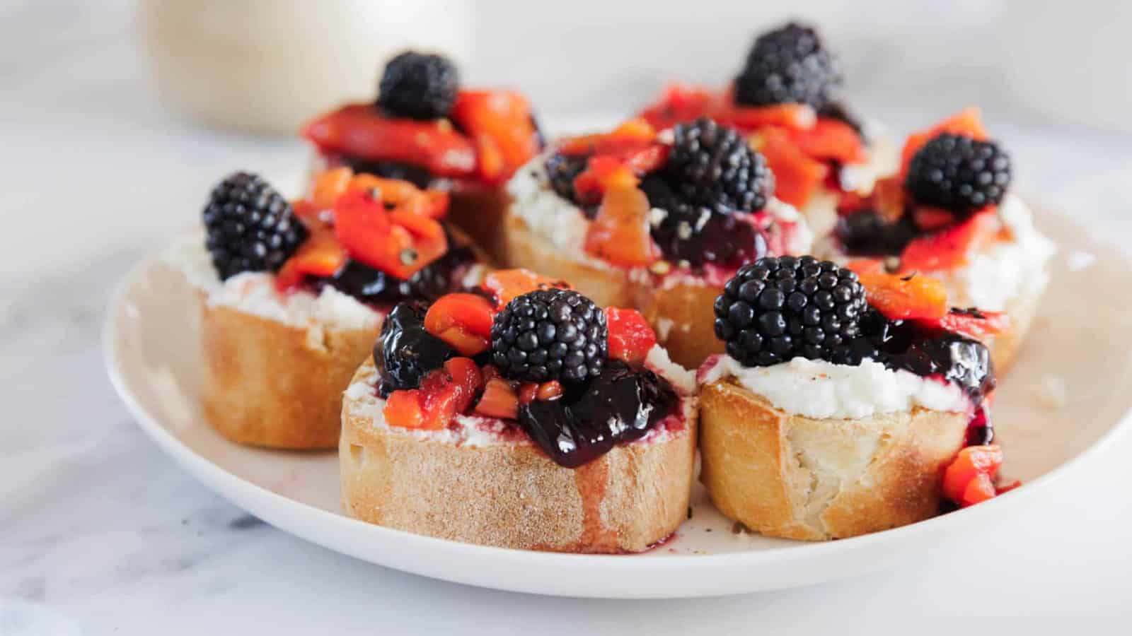 A plate of sliced baguette topped with ricotta cheese, blackberry jam, diced strawberries, and fresh blackberries, arranged on a white surface.