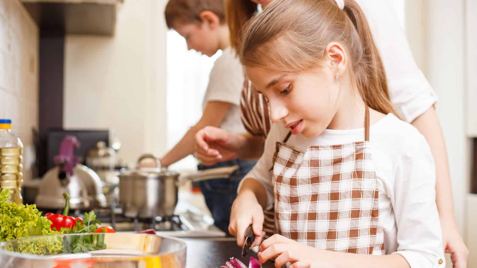 A young girl wearing a checkered apron slices a red onion on a kitchen counter. Two other people cook in the background near a stove with pots. Fresh vegetables are visible on the counter.
