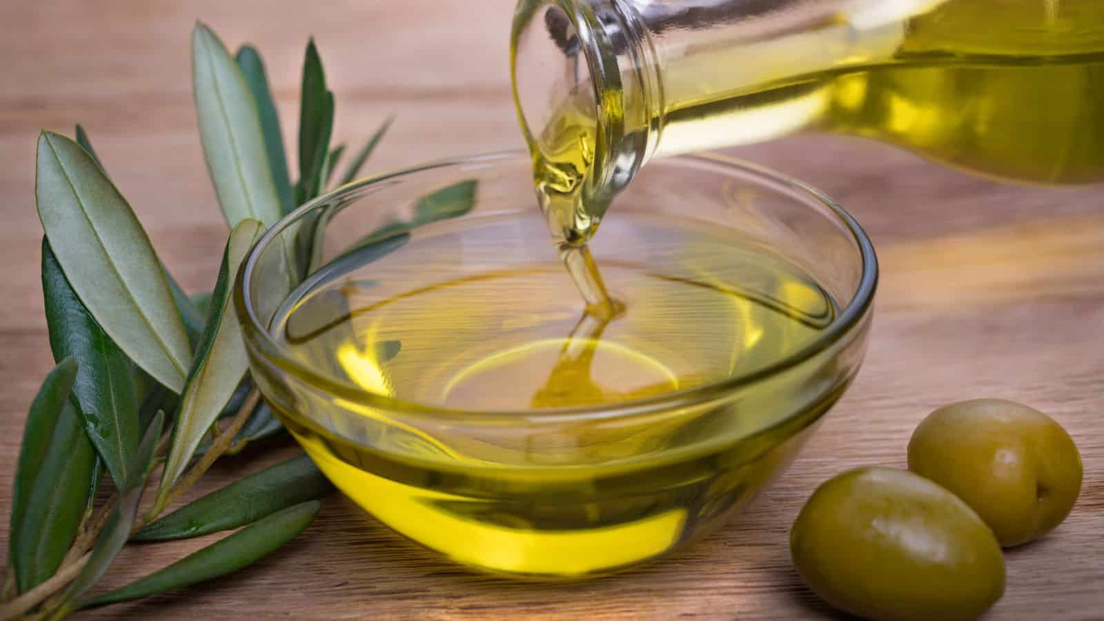 A glass bottle pours olive oil into a glass bowl, with two green olives and olive leaves resting on a wooden surface nearby.