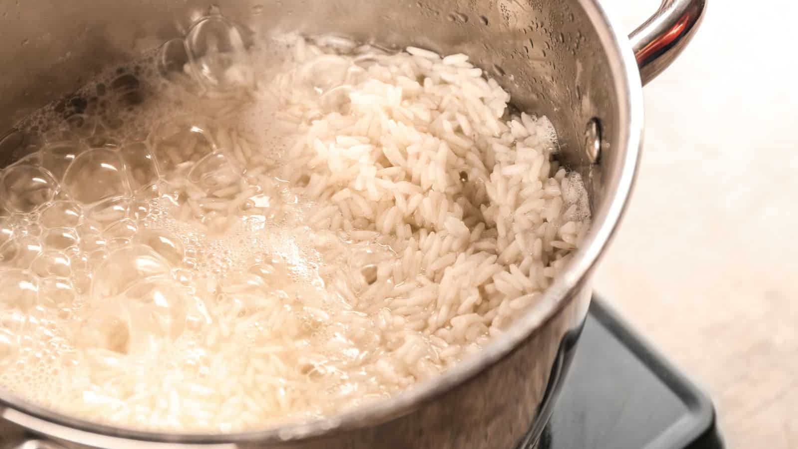 A pot of white rice is boiling on a stovetop. The rice is partially submerged in bubbling water, and steam is rising from the pot. The pot has metal sides and a visible handle.