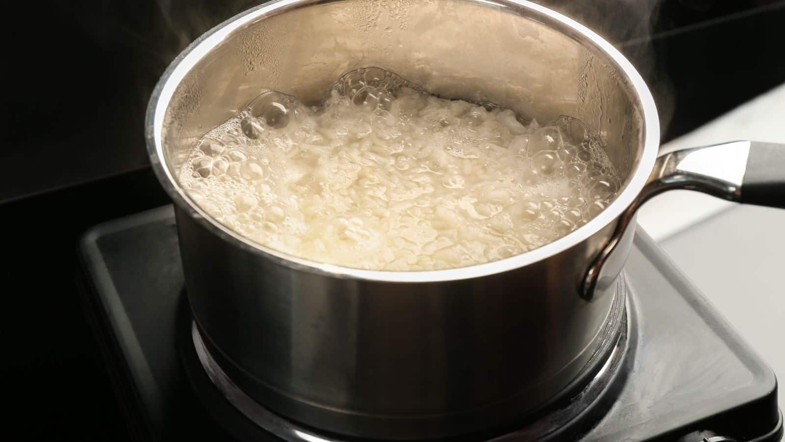 A stainless steel pot filled with boiling rice sits on an electric stove. Steam rises from the pot as bubbles form on the surface of the cooking rice. The background is dark and out of focus.