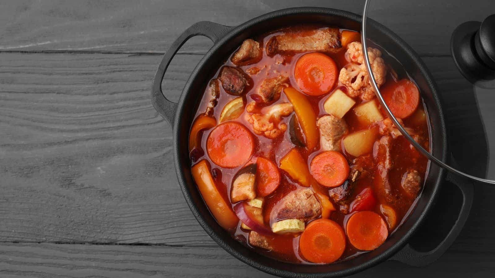 A black pot filled with a chunky stew containing carrots, potatoes, meat, and vegetables in a red broth, with a glass lid partially covering the pot, placed on a gray wooden surface.