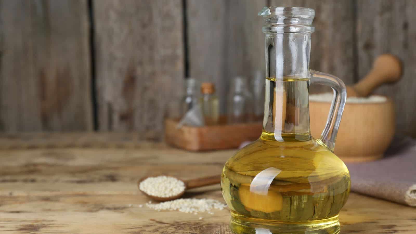 A clear glass bottle filled with light yellow oil sits on a wooden table. In the background are a wooden spoon with seeds, a wooden bowl, small glass jars, and a beige cloth, all against a rustic wooden backdrop.