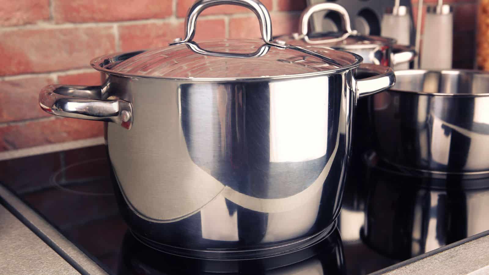 A stainless steel pot with handles and a lid sits on a black electric stovetop. There is another pot partially visible in the background. The setting includes a brick wall and kitchen utensils.