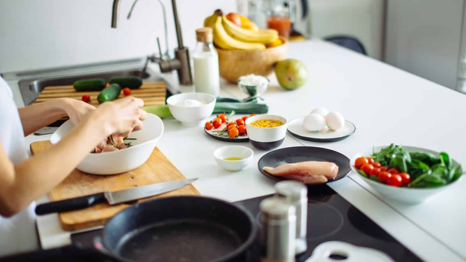 A person prepares food at a kitchen counter with ingredients including eggs, chicken breast, cheese, cherry tomatoes, spinach, and cucumbers. Various kitchen utensils and fresh fruit are also visible.