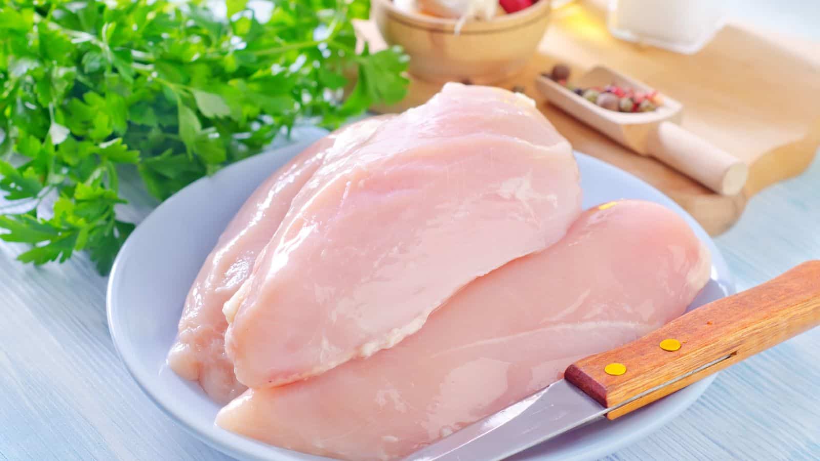 Two raw chicken breasts on a white plate with a knife, fresh parsley, and various kitchen ingredients in the background on a light-colored surface.