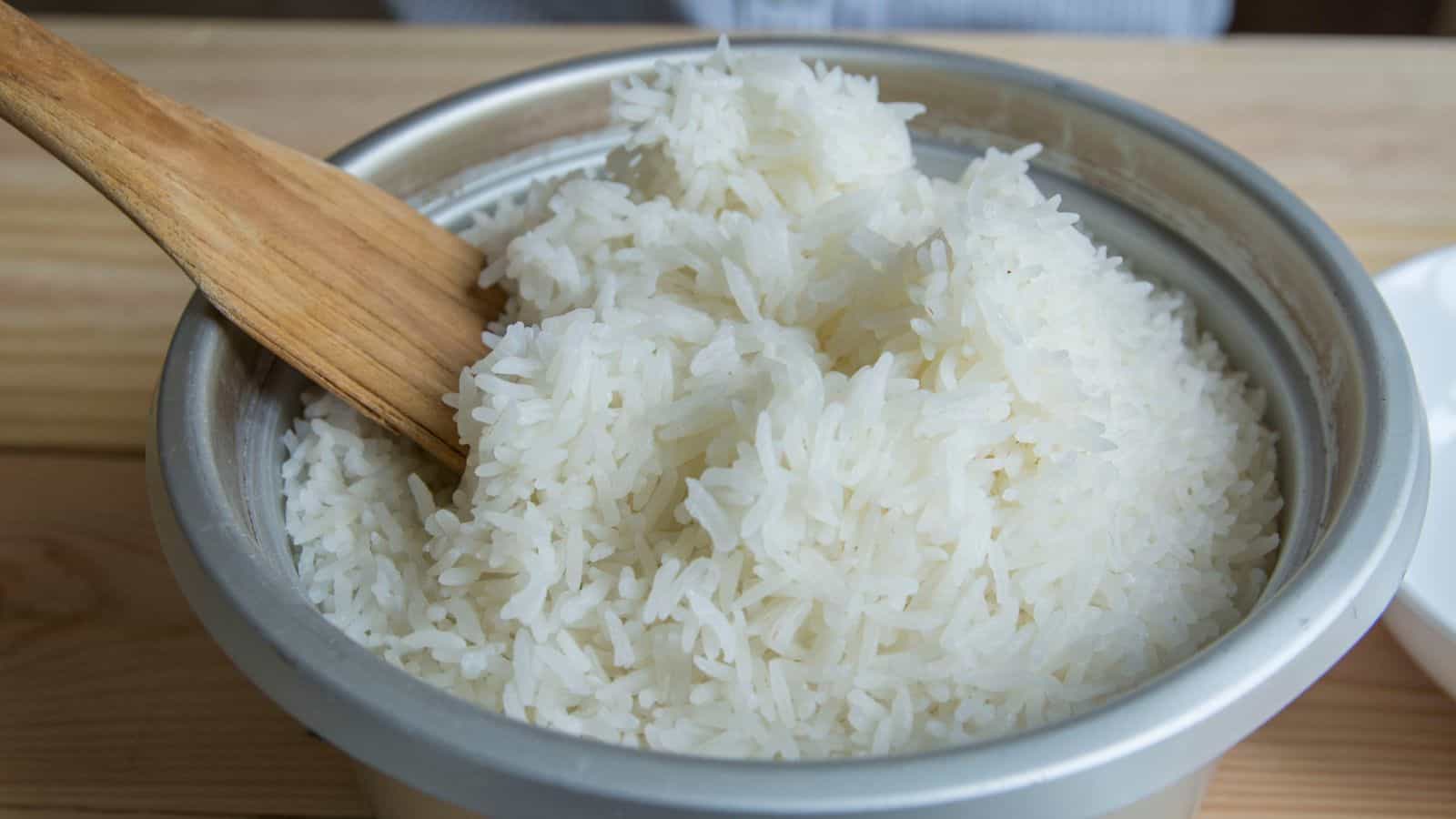 A large metal bowl filled with cooked white rice sits on a wooden table. A wooden spatula rests inside the bowl, partially mixing the rice.