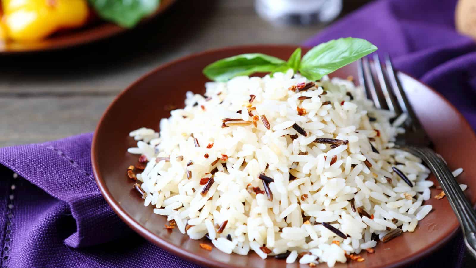 A brown plate with a serving of cooked rice garnished with herbs sits on a purple napkin. A fork and knife rest on the plate. Some grains of wild rice are mixed in with the white rice.