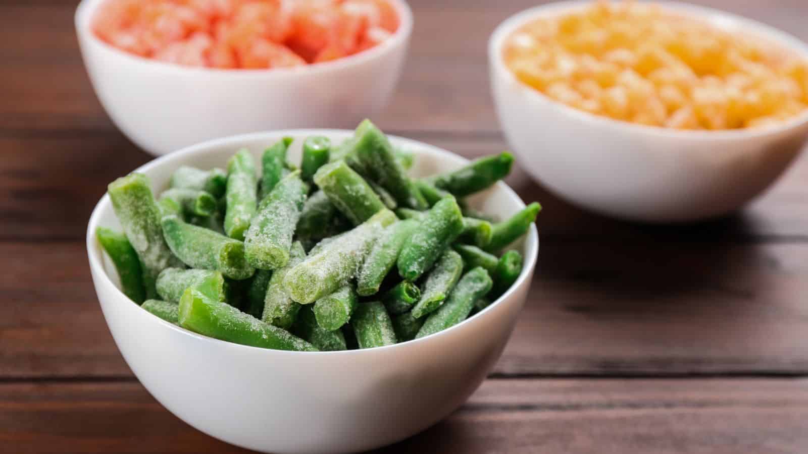 A white bowl filled with frozen green beans in the foreground, with two other white bowls containing chopped vegetables, possibly carrots and tomatoes, in the background on a wooden table.