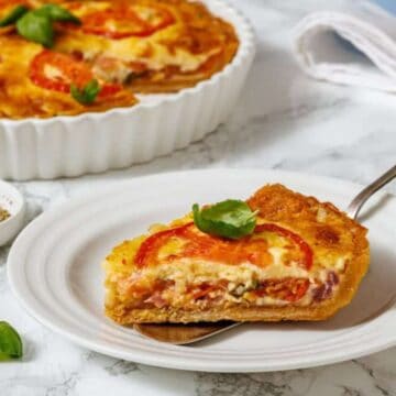 A slice of tomato and cheese quiche on a white plate with a fork, garnished with fresh basil. The whole quiche is visible in a baking dish behind it, along with a small bowl of pepper and basil leaves on the table.