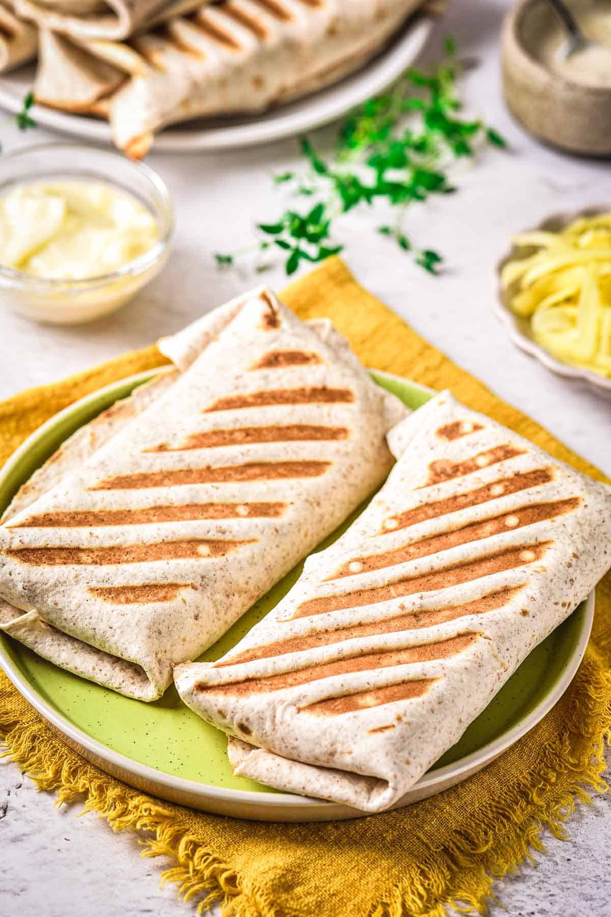 Two grilled wraps with visible grill marks are placed on a green plate atop a yellow cloth. In the background, a bowl of sauce, sliced vegetables, and additional wraps are visible.