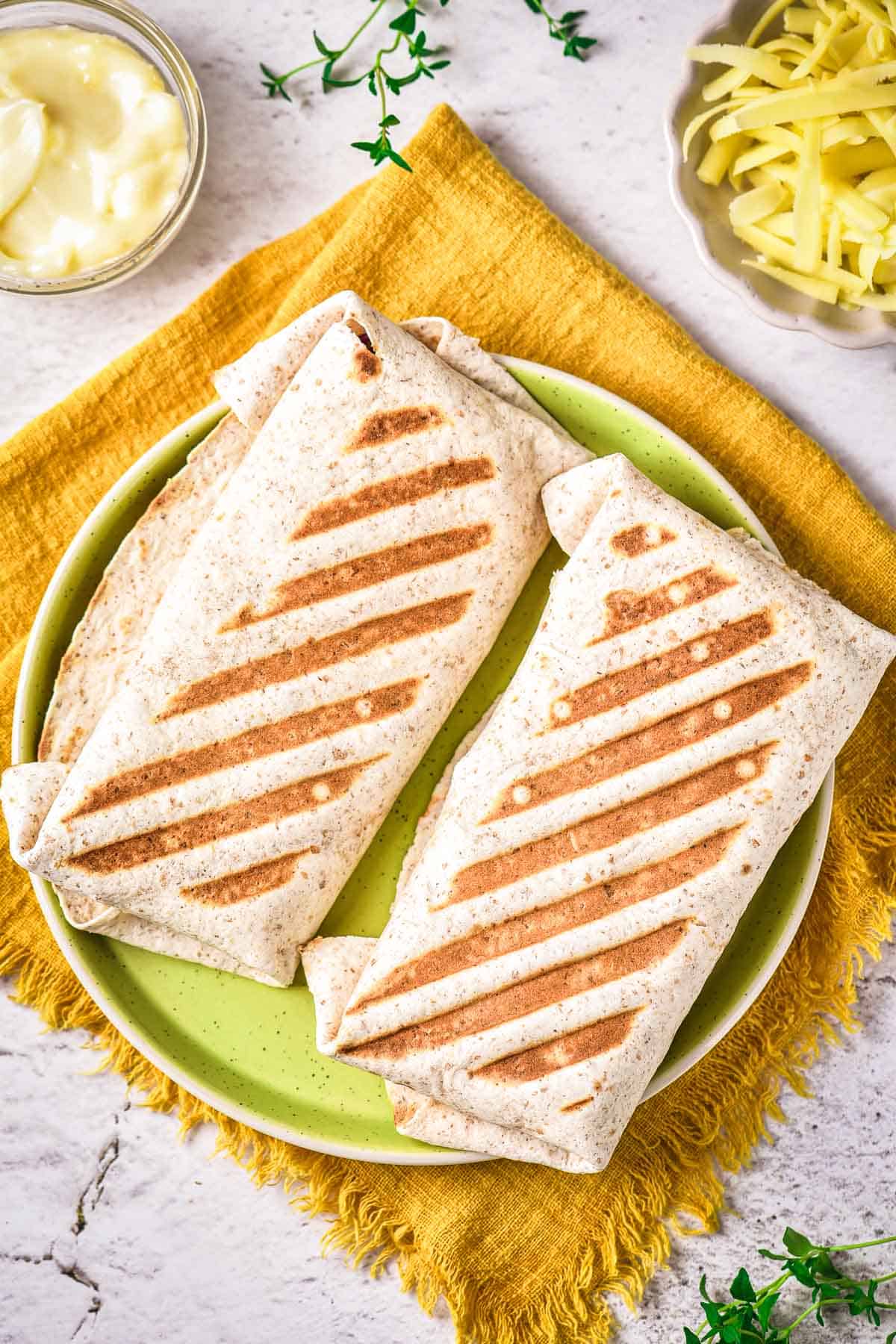 Two grilled wraps with visible grill marks are placed side by side on a green plate over a yellow napkin. Sliced cheese and a small bowl of mayonnaise are nearby on a white surface.