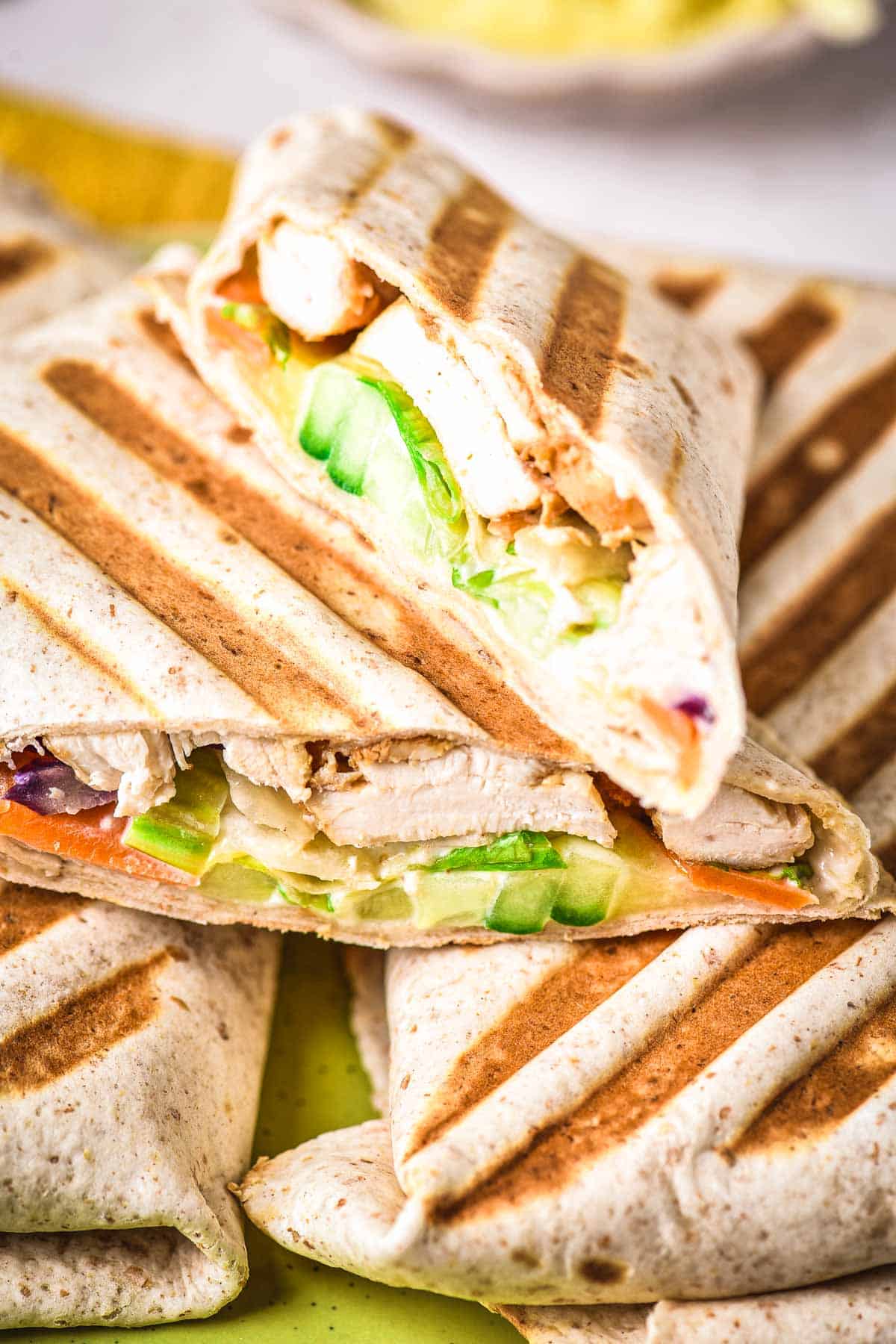 A close-up of grilled wraps cut in half, revealing layers of sliced chicken, lettuce, cucumber, and other vegetables inside a toasted tortilla. The wraps are stacked on top of each other.