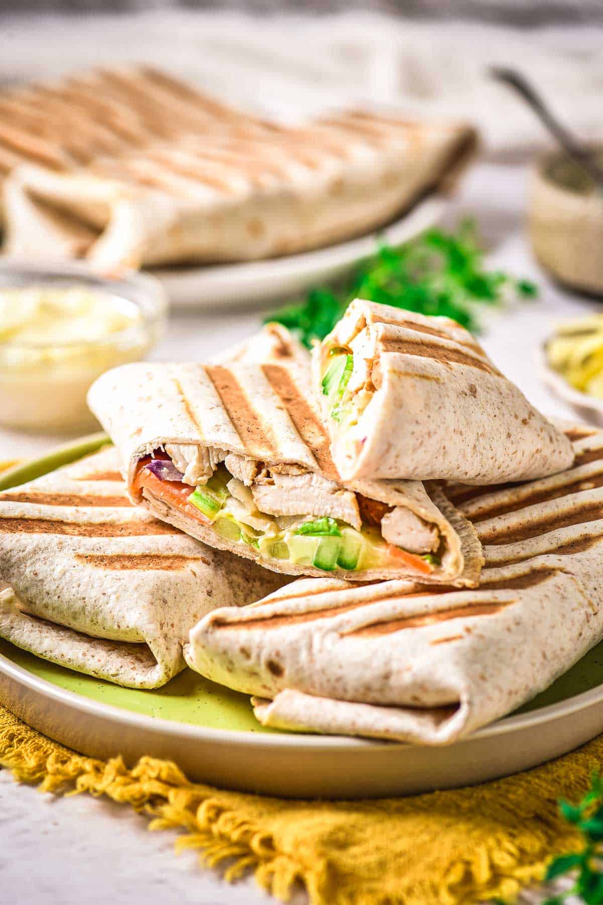 Three grilled wraps cut in half and stacked on a green plate, showing fillings of chicken, lettuce, tomato, and other vegetables. More wraps and a bowl of sauce are in the background on a white table.