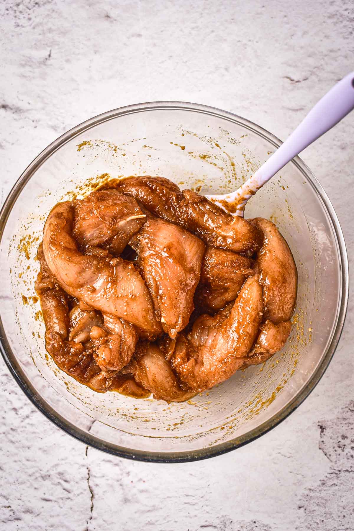 Raw chicken strips coated in a brown marinade sit in a clear glass bowl with a white spoon, on a light textured surface.