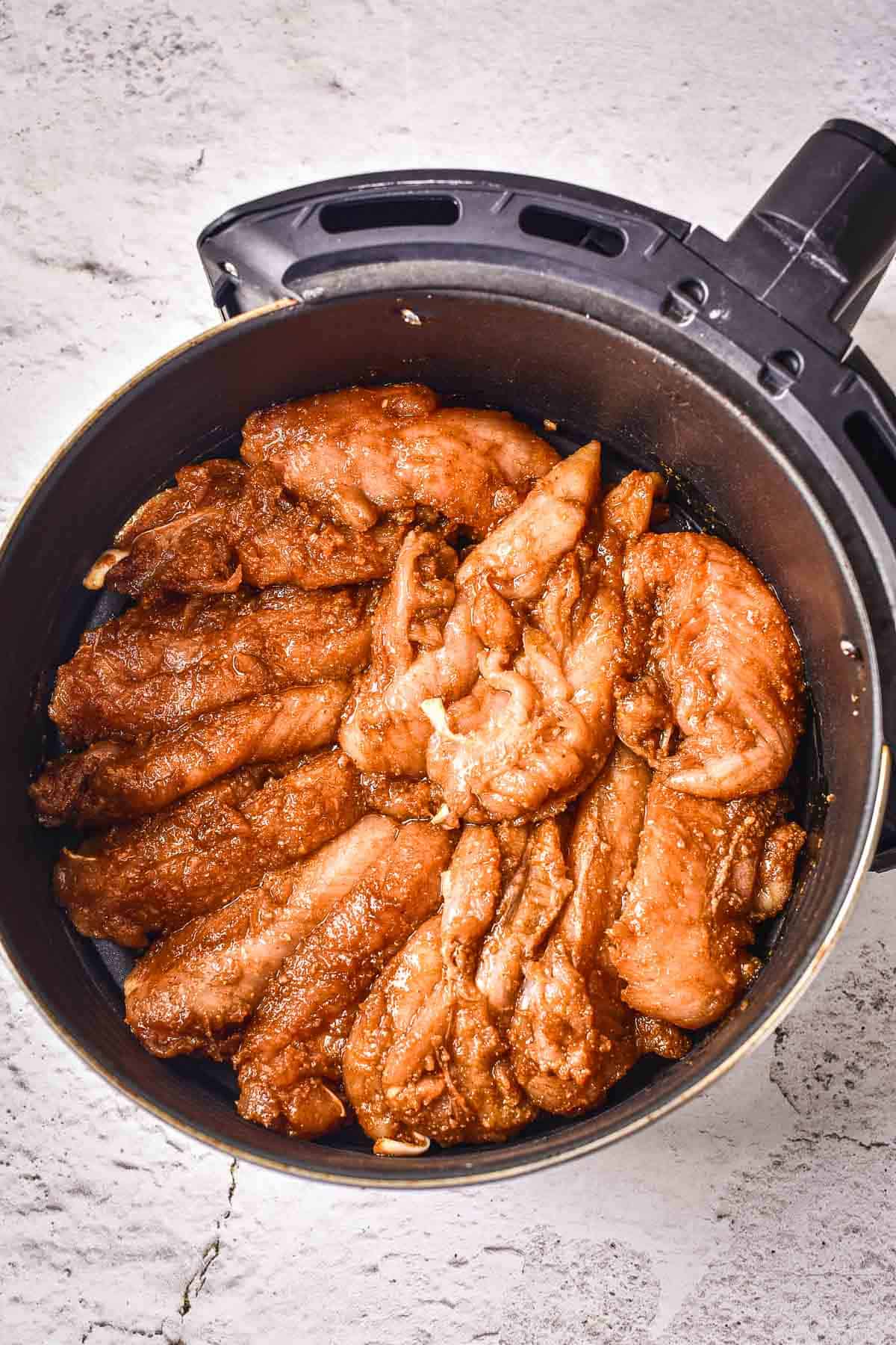 Raw, marinated chicken strips arranged in a black air fryer basket on a light textured surface, ready to be cooked.