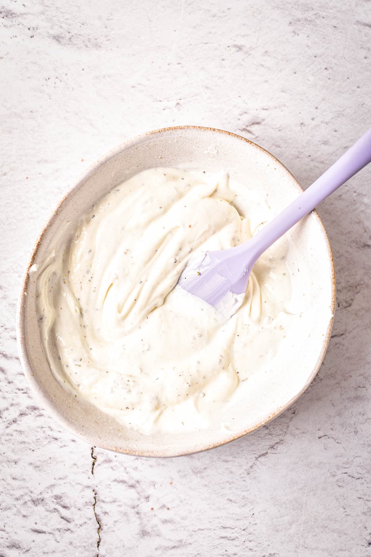 A bowl filled with a creamy white sauce or dip, speckled with herbs, sits on a light textured surface. A light purple spatula rests in the bowl with some sauce on it.