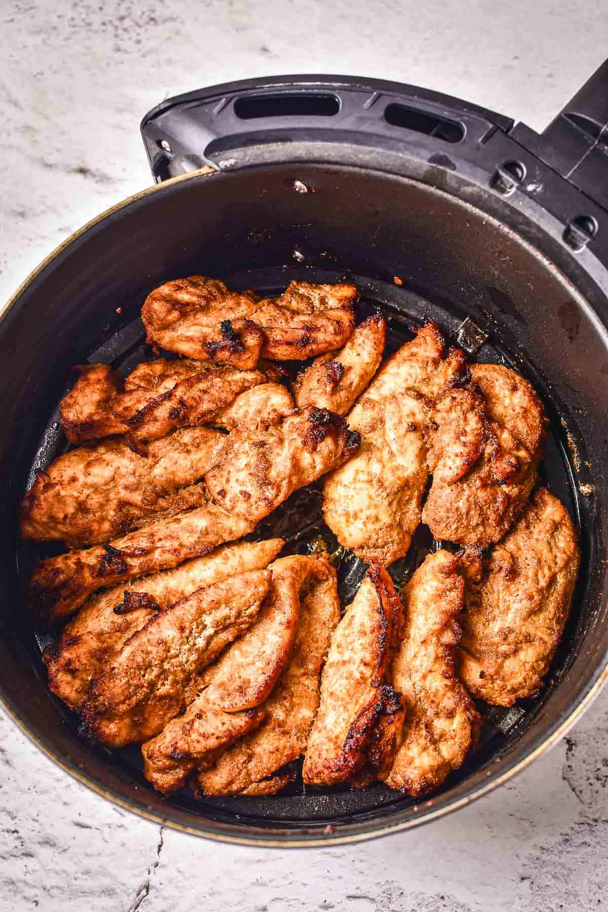 Several cooked, seasoned chicken tenders are arranged inside the basket of an air fryer on a white textured surface.