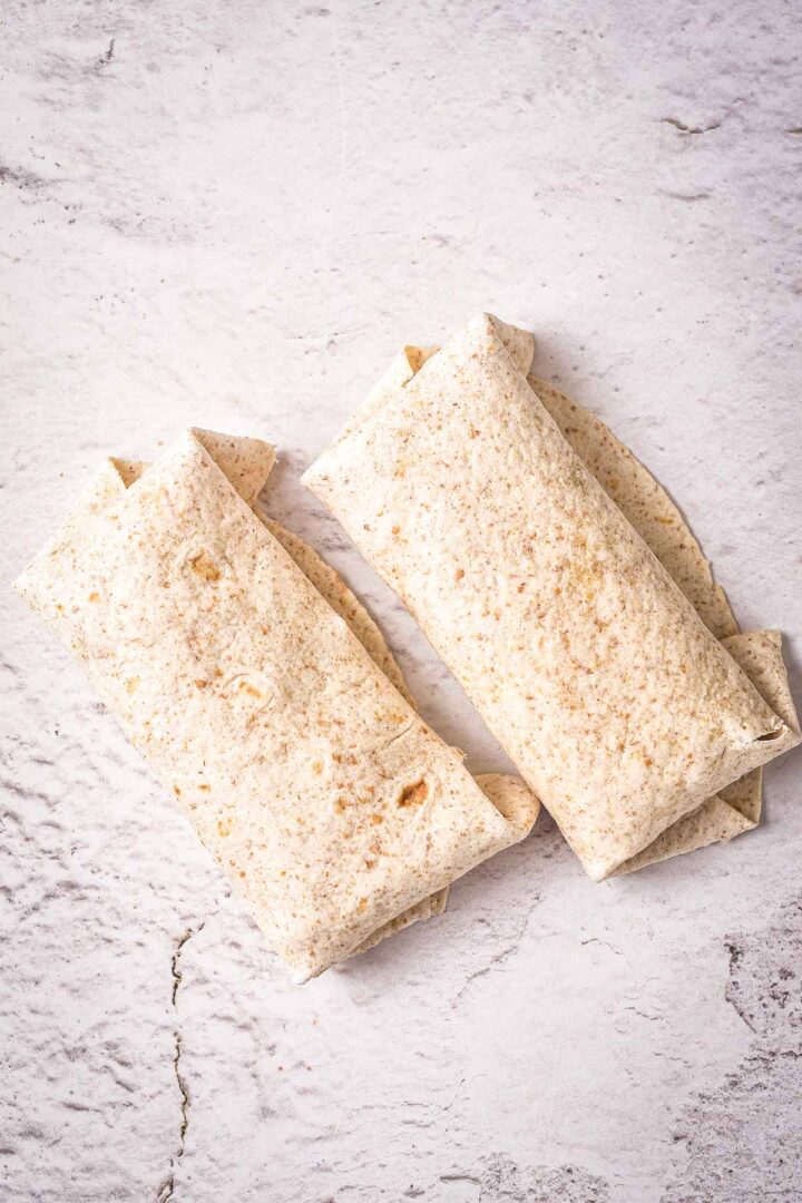 Two whole wheat tortillas are folded into rectangular wraps and placed side by side on a textured light-colored surface.