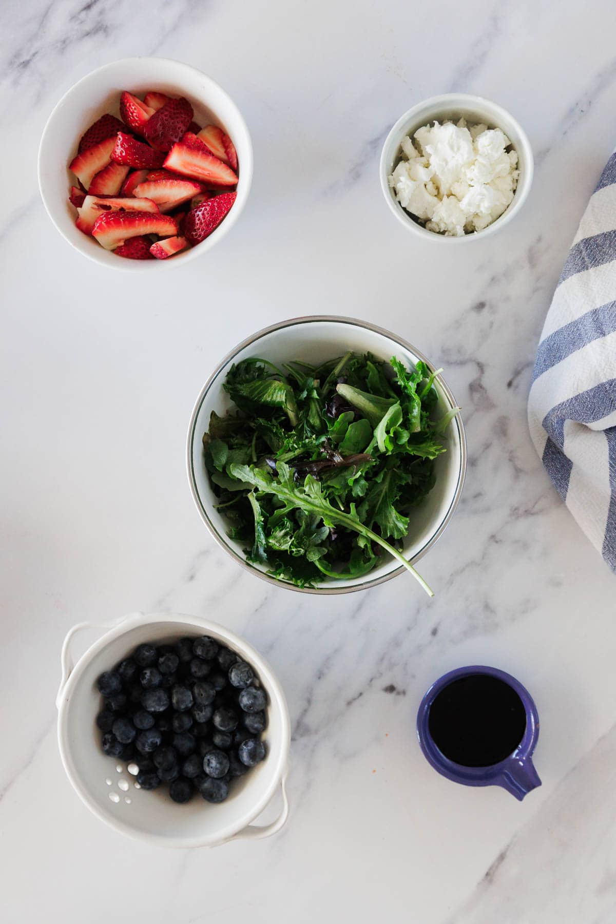 A bowl of mixed salad greens is surrounded by bowls of sliced strawberries, blueberries, crumbled cheese, and a small cup of dark dressing on a white marble surface with a blue and white striped cloth.