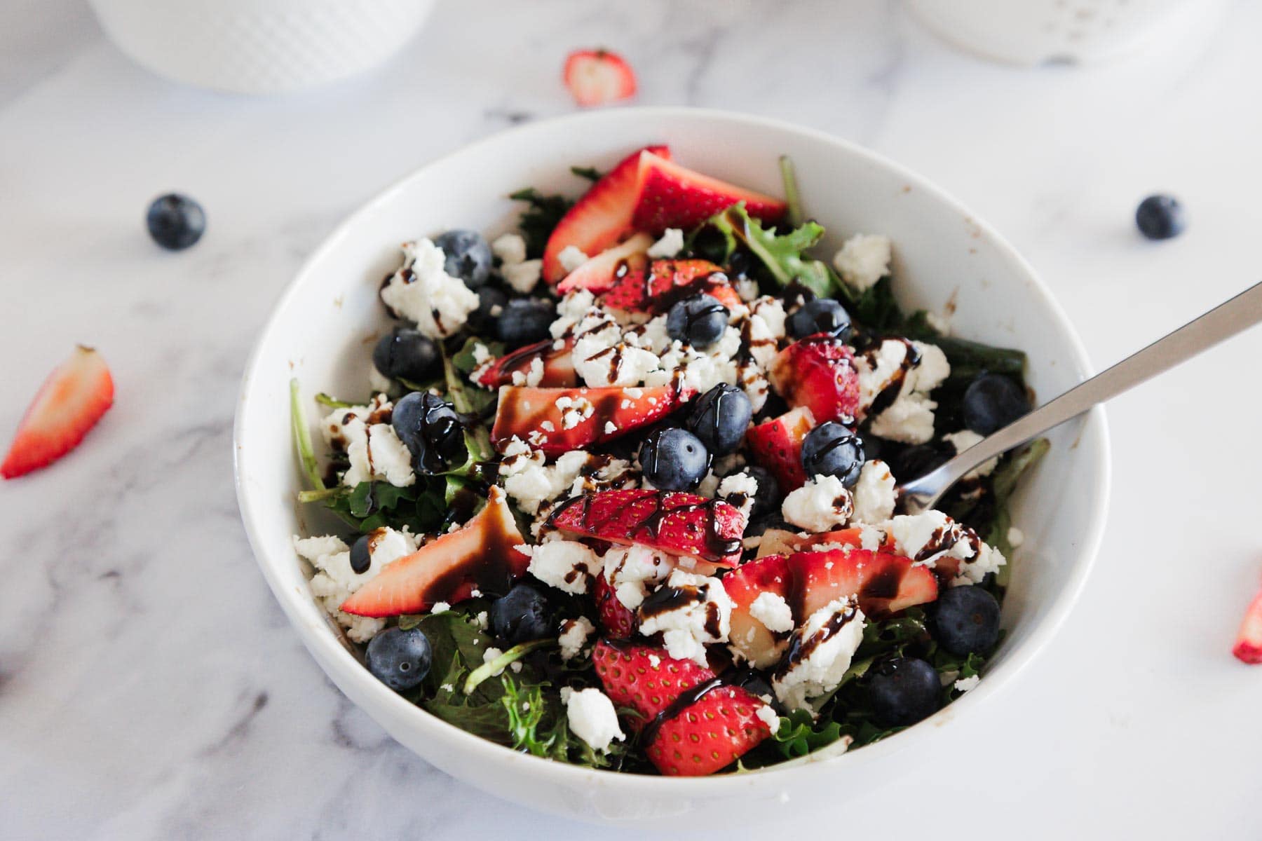 A white bowl filled with a salad of arugula, sliced strawberries, blueberries, crumbled feta cheese, and a drizzle of balsamic glaze. A spoon rests in the bowl, with berries and salad scattered on the table.