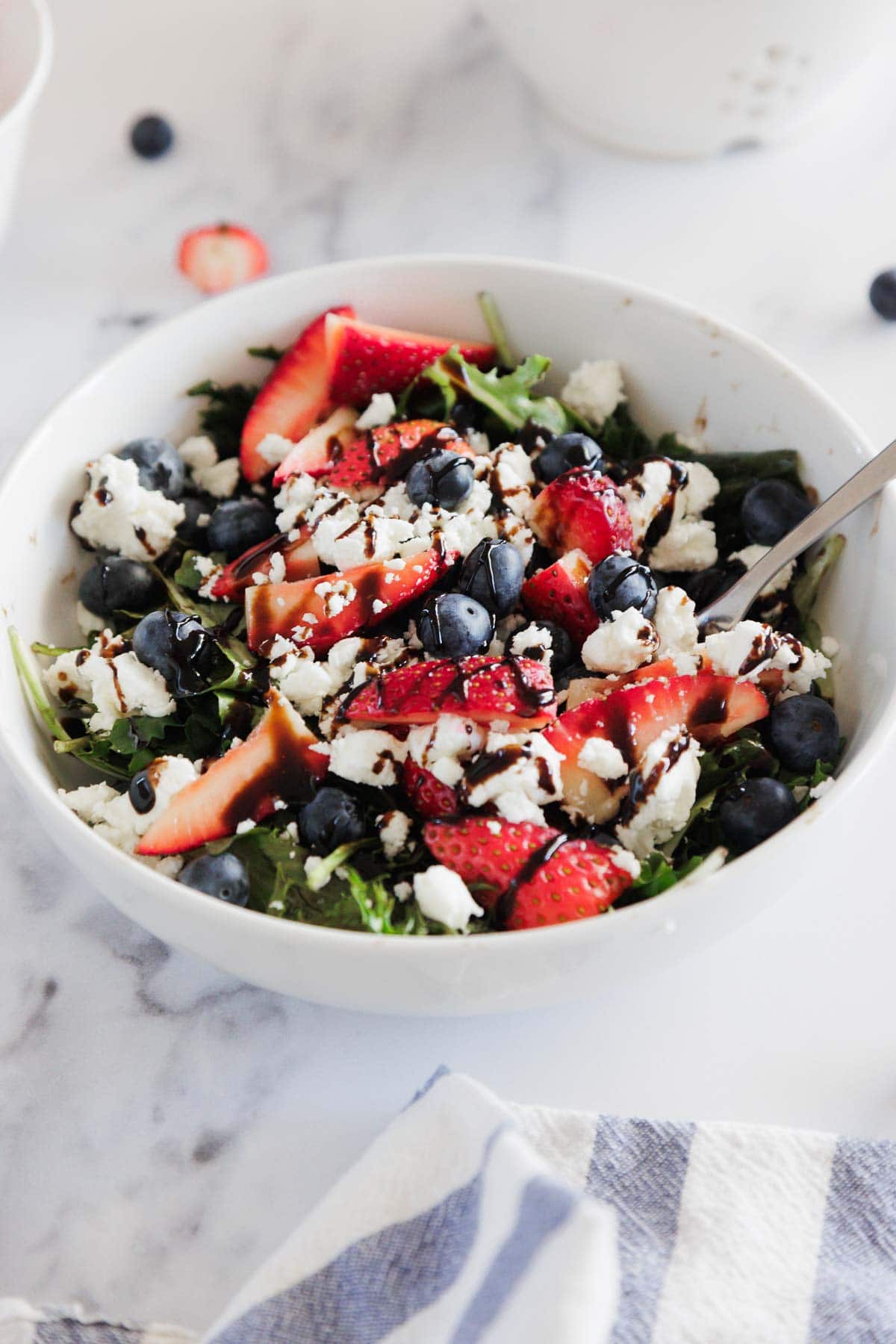 A white bowl filled with a salad of arugula, sliced strawberries, blueberries, crumbled feta cheese, and a drizzle of balsamic glaze. A fork rests in the bowl, which sits on a marble surface.