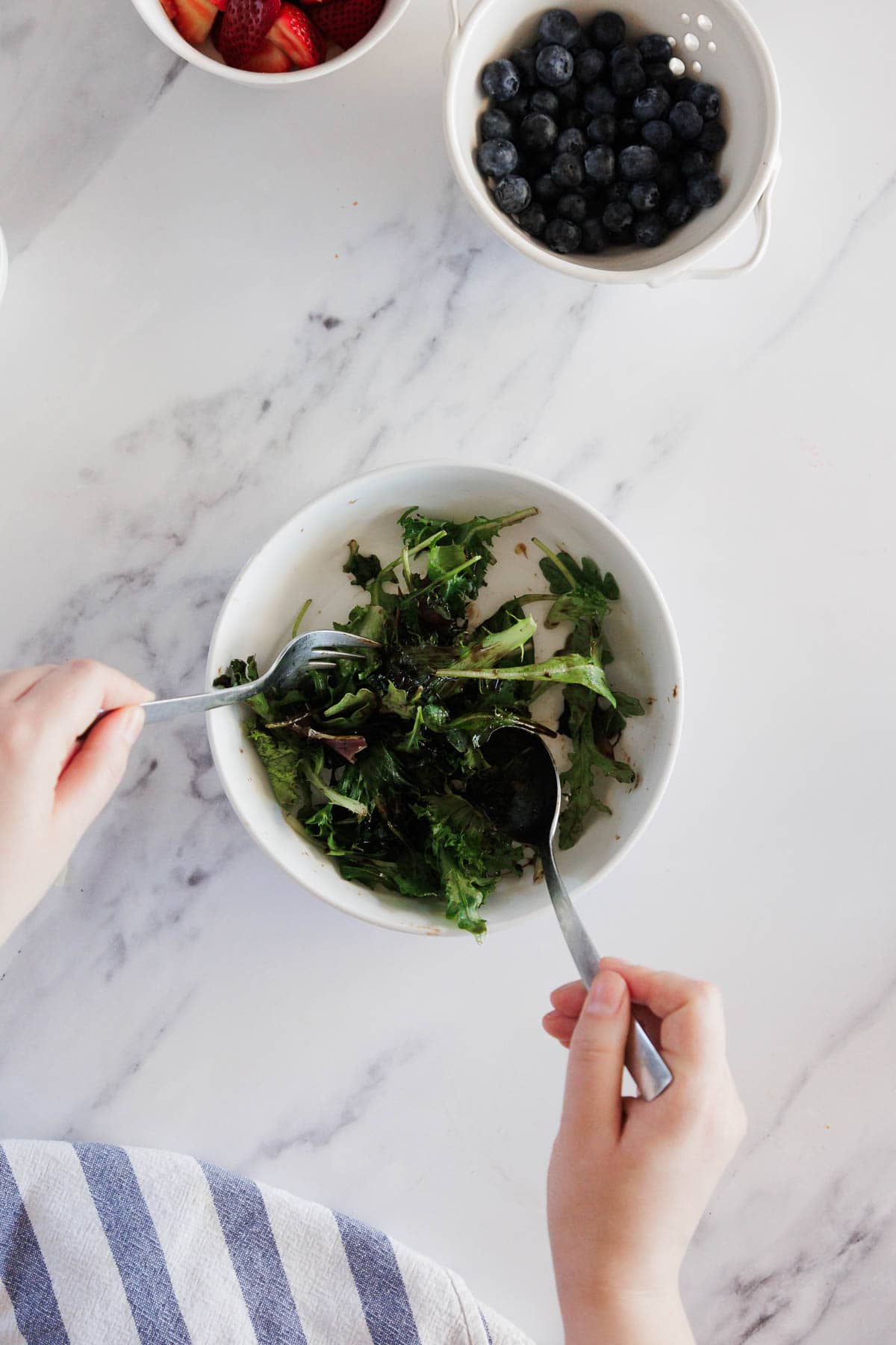 A person mixes a green salad in a white bowl with two forks. Nearby are bowls of blueberries and sliced strawberries on a white marble surface with a striped towel partially visible.