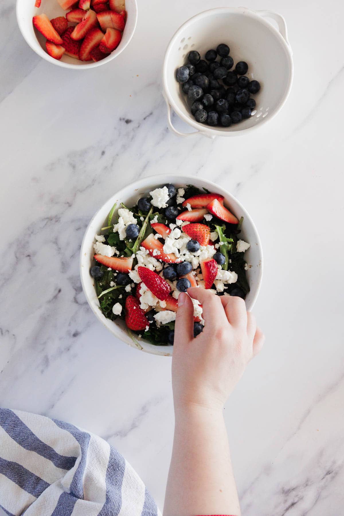 A hand reaches into a bowl of salad containing strawberries, blueberries, and crumbled white cheese, with extra strawberries and blueberries in separate bowls nearby on a marble surface.