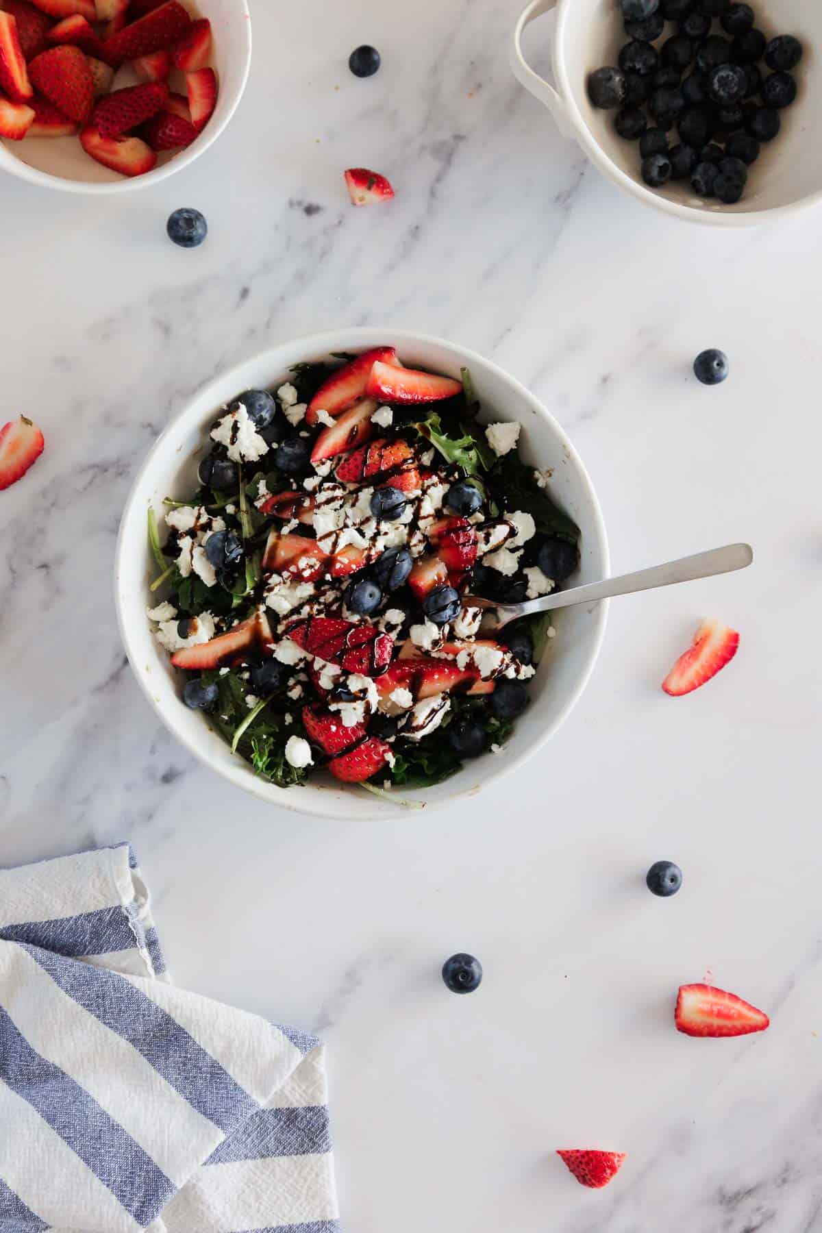 A white bowl filled with a salad of leafy greens, blueberries, strawberries, crumbled cheese, and a drizzle of dressing sits on a marble surface with a spoon. Fresh blueberries and strawberries are in small bowls nearby.