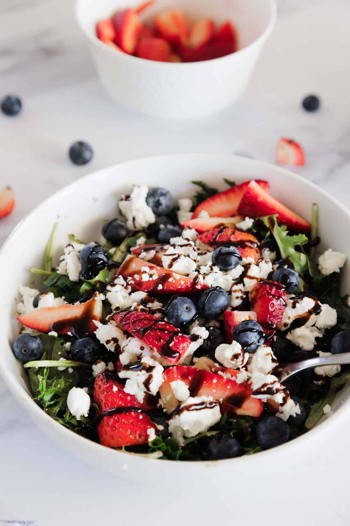 A white bowl filled with a salad made of arugula, sliced strawberries, blueberries, crumbled feta cheese, and balsamic glaze. In the background, a small bowl contains more sliced strawberries.