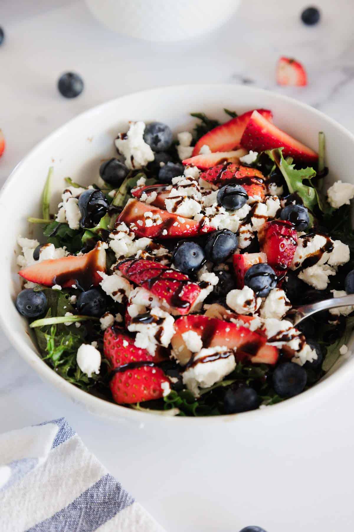 A white bowl filled with a salad containing arugula, sliced strawberries, blueberries, crumbled feta cheese, and a drizzle of balsamic glaze, with a spoon in the bowl and additional berries on the table nearby.
